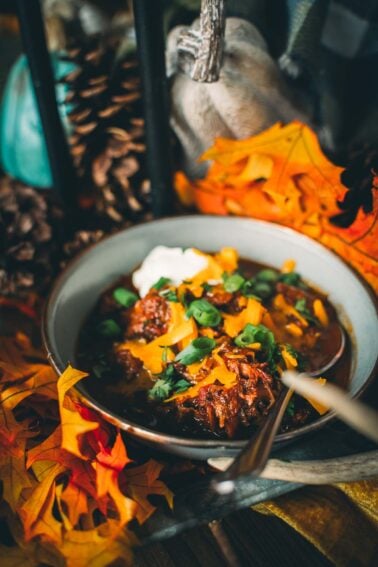 A bowl of over the top chili topped with cheese, herbs, and a dollop of cream, surrounded by autumn leaves and pine cones.