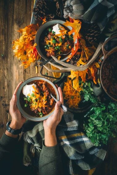 Two bowls of chili topped with sour cream, cheese, and green onions are held above a fall-themed table with leaves and a plaid blanket.