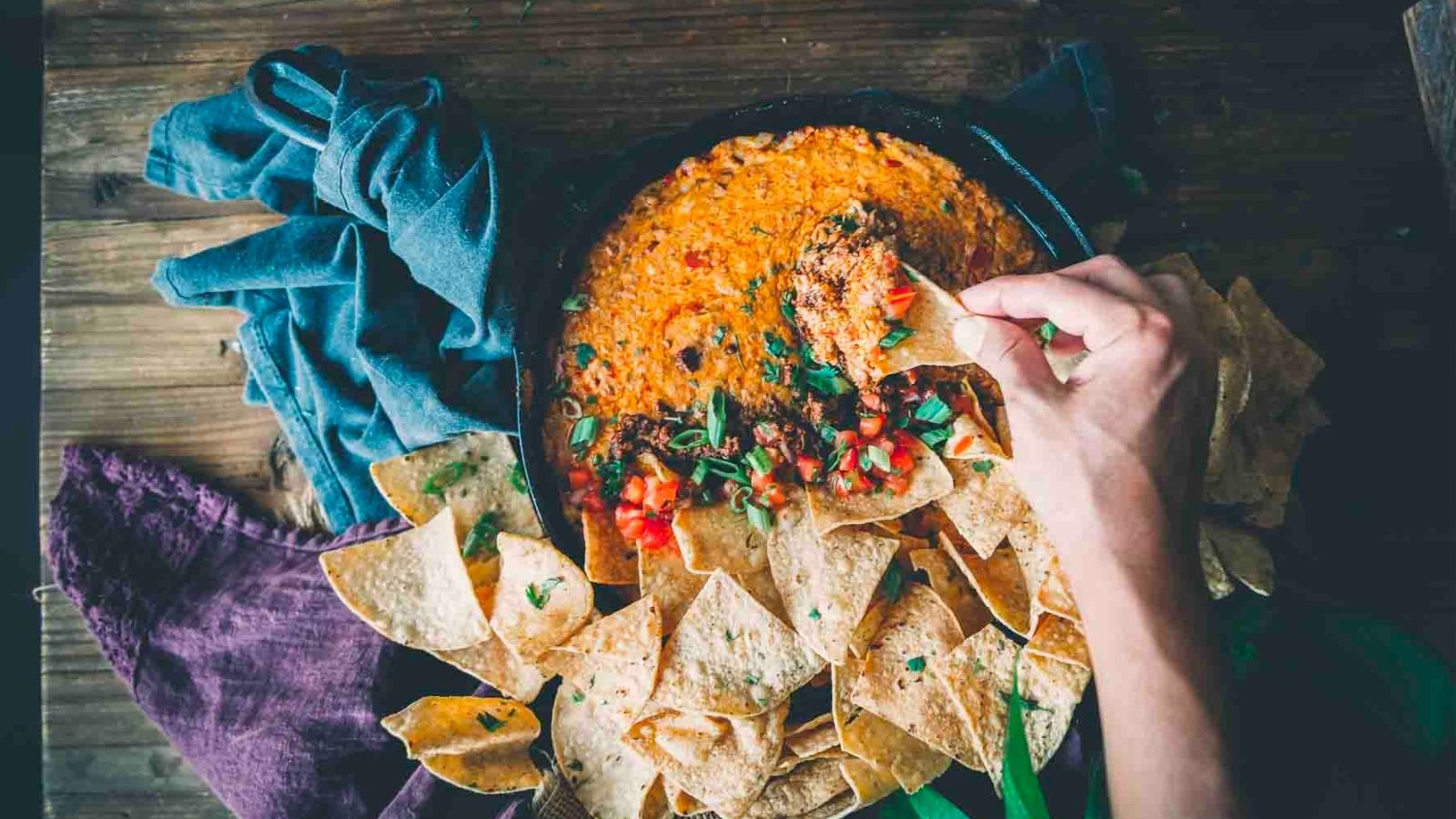 A hand dips a tortilla chip into a pan of cheesy, seasoned dip, surrounded by more chips on a wooden surface.