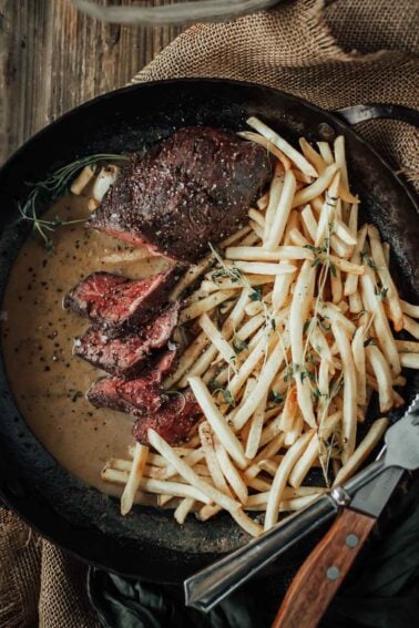 Sous vide hanger steak slices with peppercorn sauce and a pile of crispy fries in a black skillet, garnished with fresh thyme.