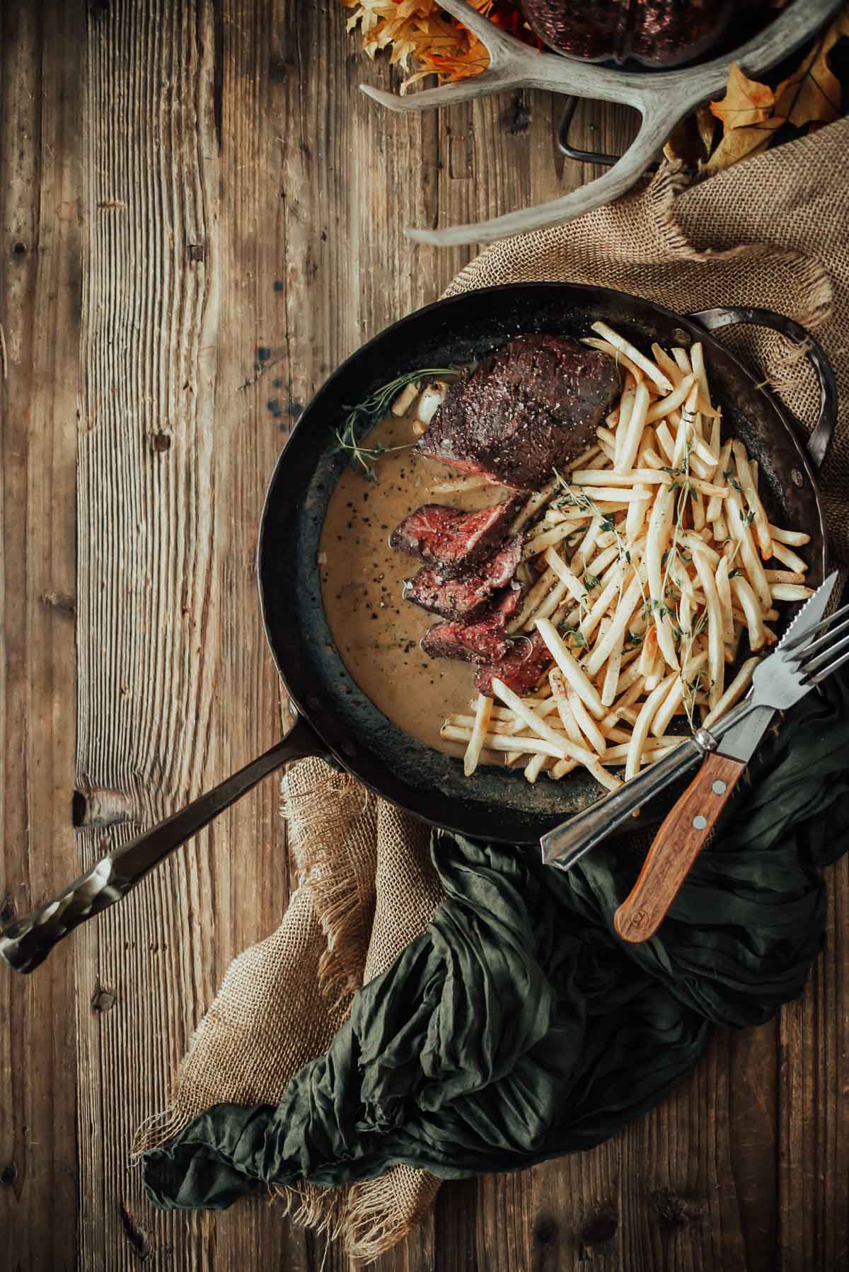 A pan of sous vide hanger steak in pepper sauce with fries, garnished with herbs. Set on a rustic wooden table with a fork and knife.