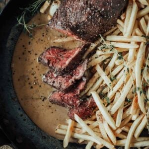 A plate with sliced sous vide hanger steak, thin fries, and creamy sauce, garnished with herbs.