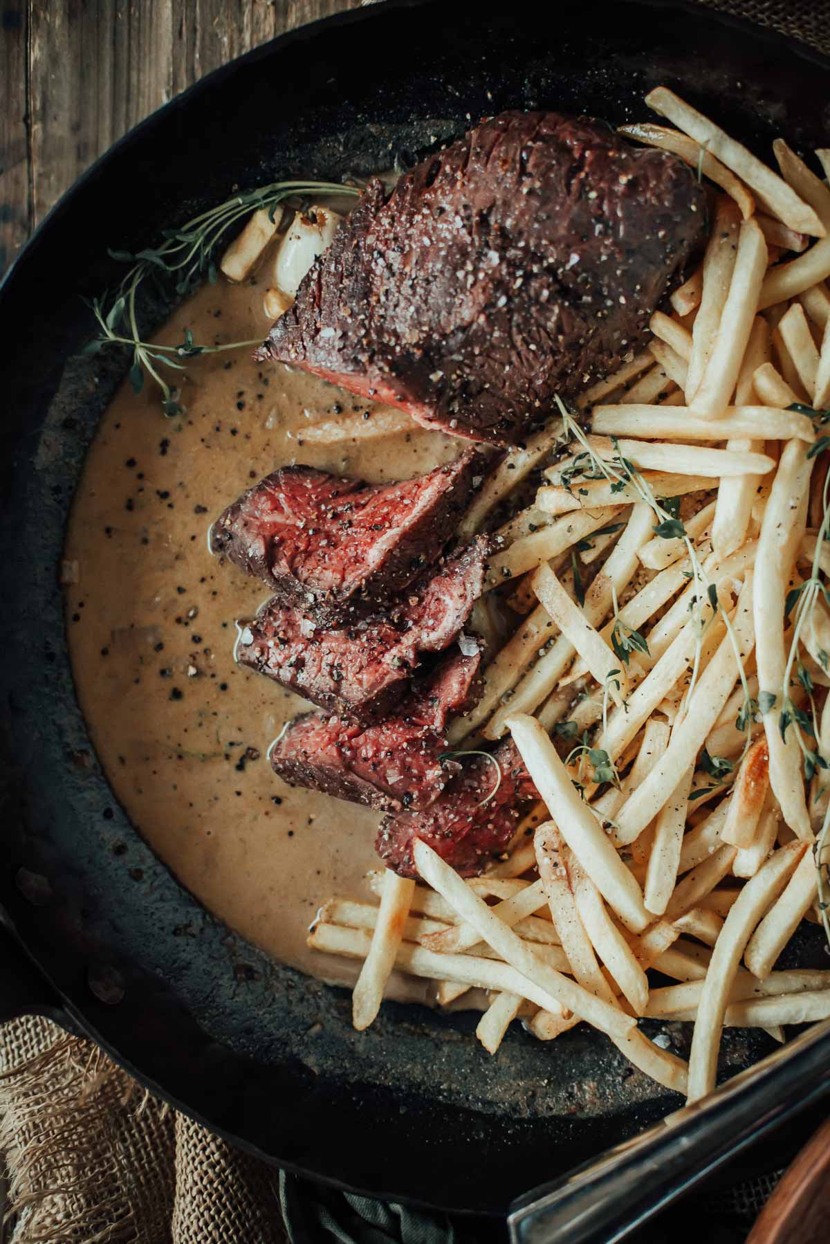 A plate with sliced sous vide hanger steak, thin fries, and creamy sauce, garnished with herbs.