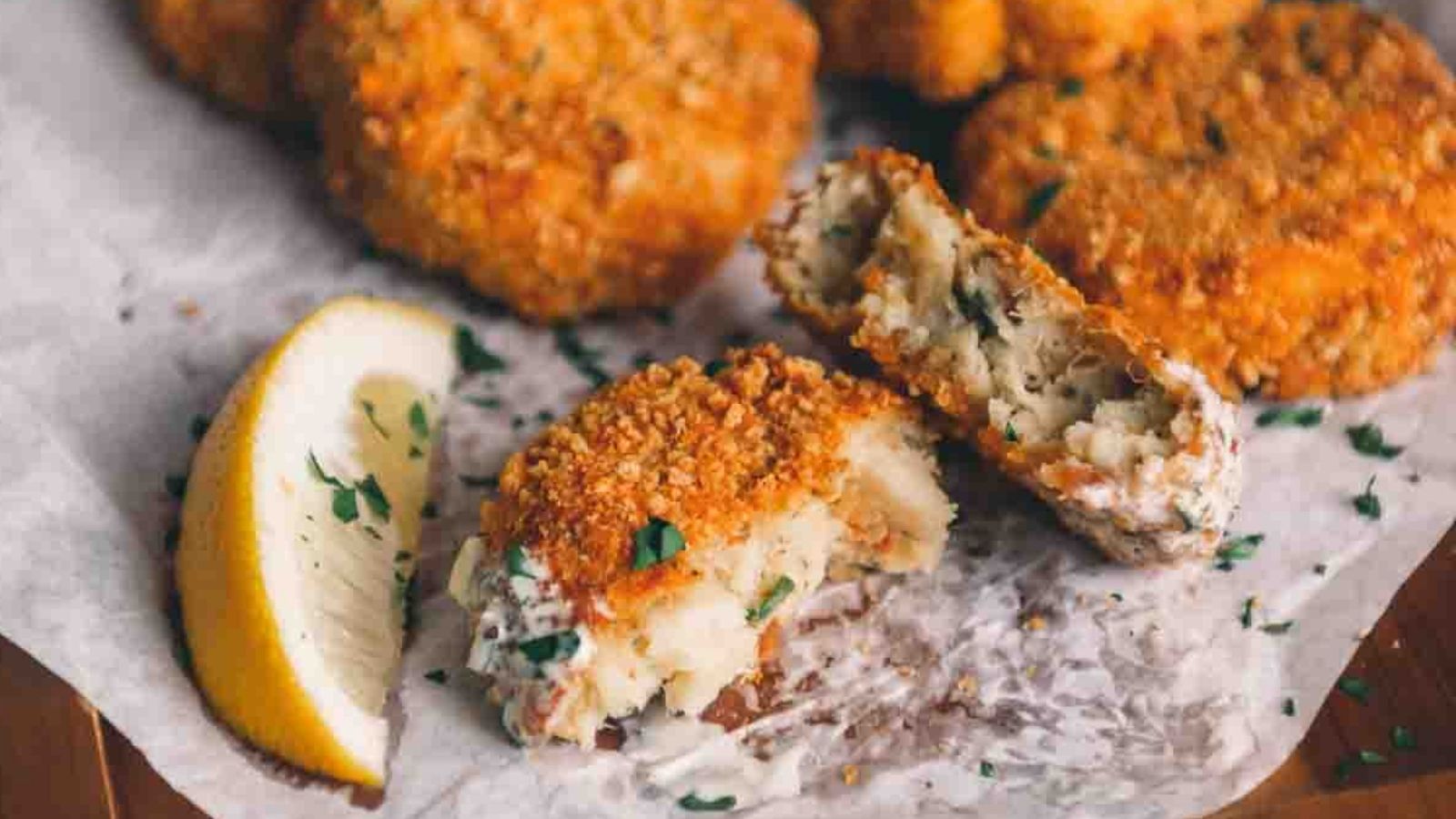 Fried crab cakes on parchment paper with a lemon wedge. One cake is broken, revealing its soft interior. Parsley garnish is visible.