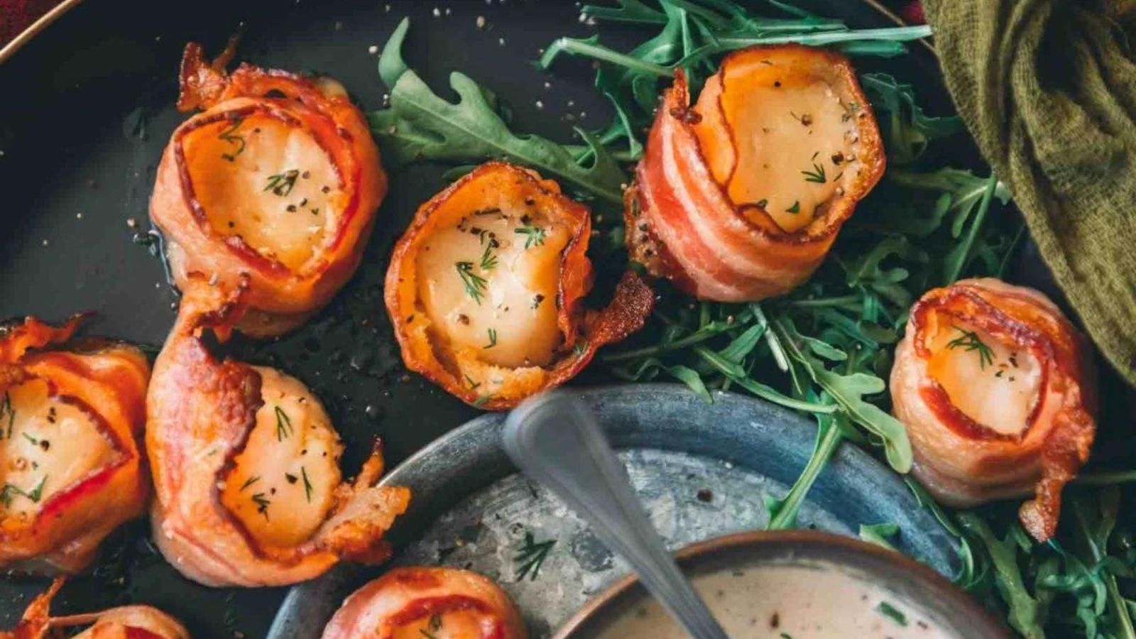 Bacon-wrapped scallops garnished with herbs on arugula leaves, served with a bowl of creamy dipping sauce.