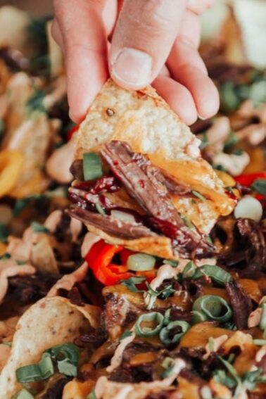 A hand holding a nacho chip topped with sliced meat, cheese, and vegetables, with a tray of nachos underneath.