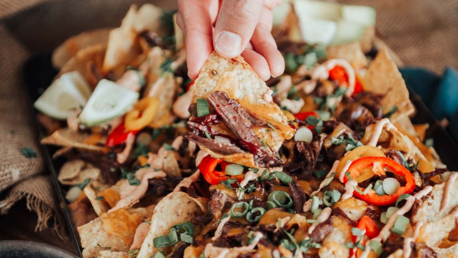 A hand holding a nacho chip topped with sliced meat, cheese, and vegetables, with a tray of nachos underneath.