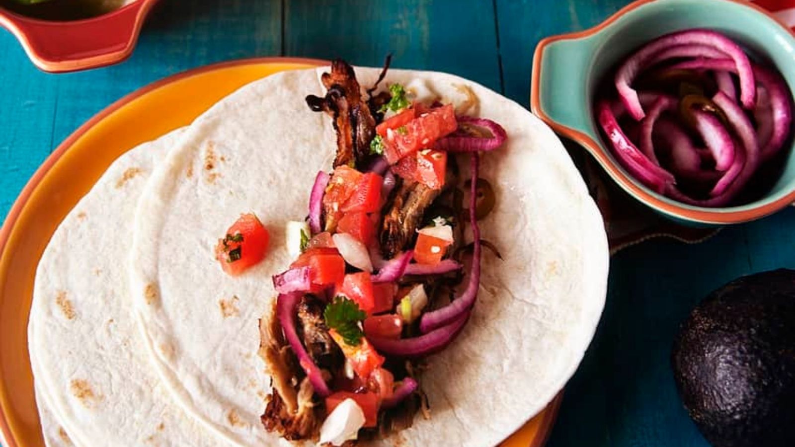 Two tortillas with meat, chopped tomatoes, onions, and herbs, on an orange plate. A bowl with sliced onions and an avocado are nearby on a blue table.