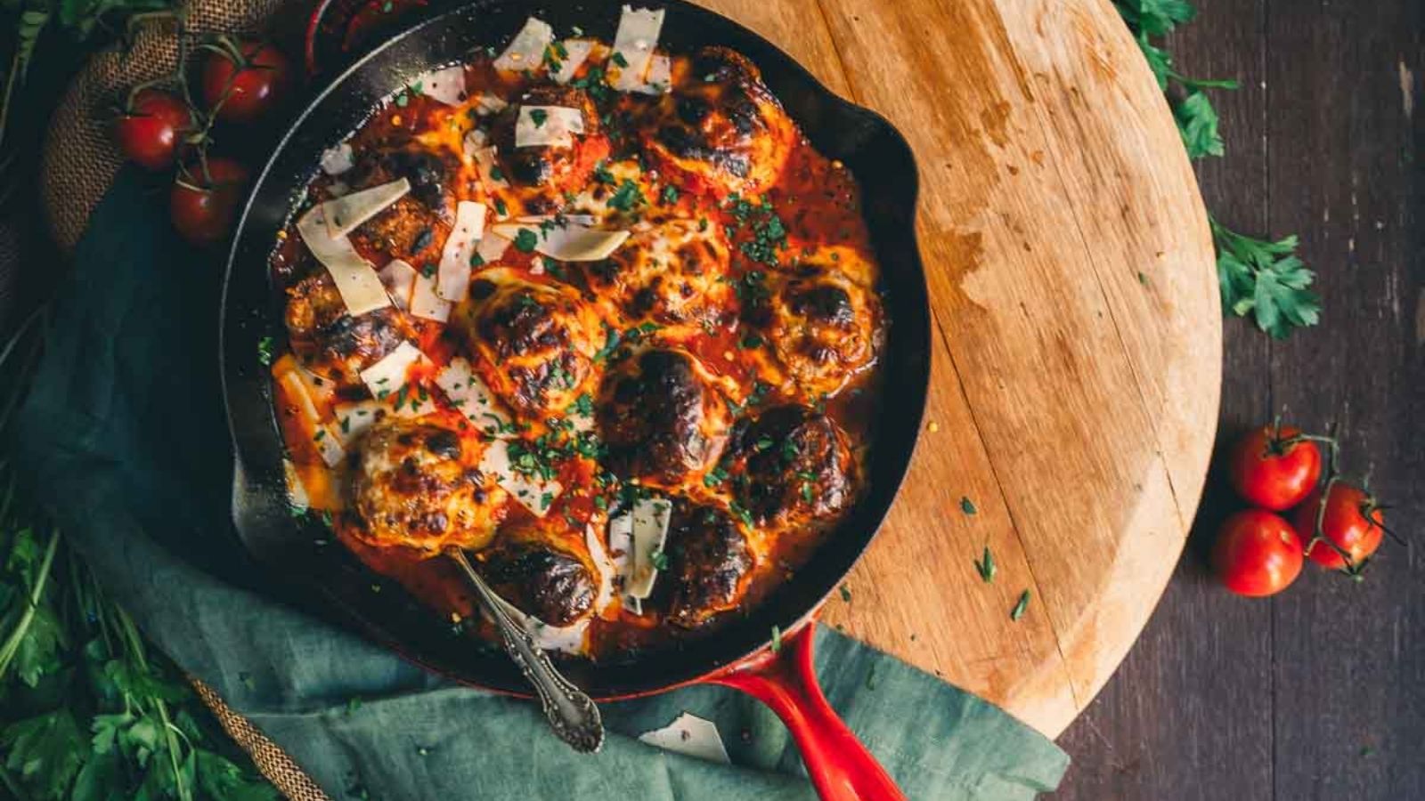 A skillet filled with meatballs in tomato sauce, garnished with parsley and Parmesan cheese, placed on a round wooden board with cherry tomatoes and fresh herbs nearby.