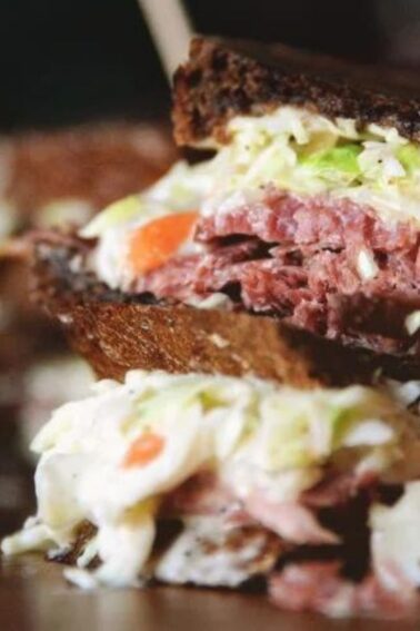Close-up of a sandwich with rye bread, corned beef, coleslaw, and dressing, skewered and served on a wooden surface.