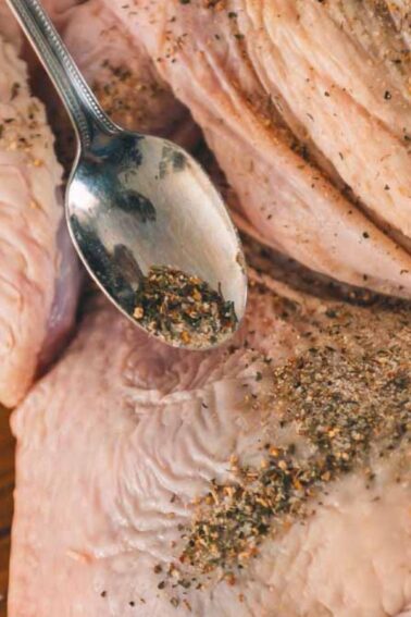 A raw turkey being seasoned with spices using a spoon on a wooden surface.