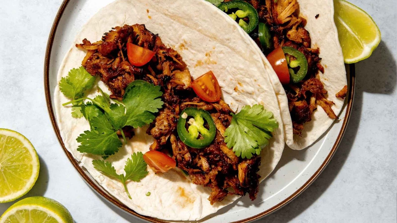 Two tacos with shredded meat, sliced jalapeños, cherry tomatoes, and cilantro on a plate. Lime wedges are nearby.