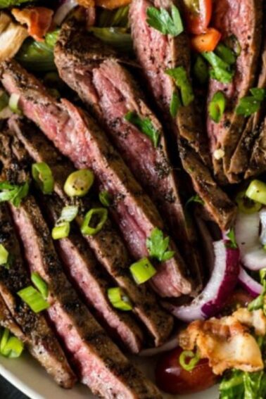 Sliced grilled steak served on a plate with a salad of lettuce, onions, tomatoes, mushrooms, and bell peppers. Garnished with green onions and herbs. Knife and cloth nearby.