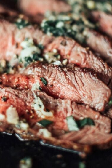 Close-up of sliced medium-rare steak topped with garlic and herbs on a dark surface.