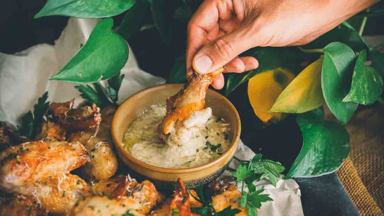 A hand dips a chicken wing into a bowl of creamy sauce, surrounded by parsley and foliage.