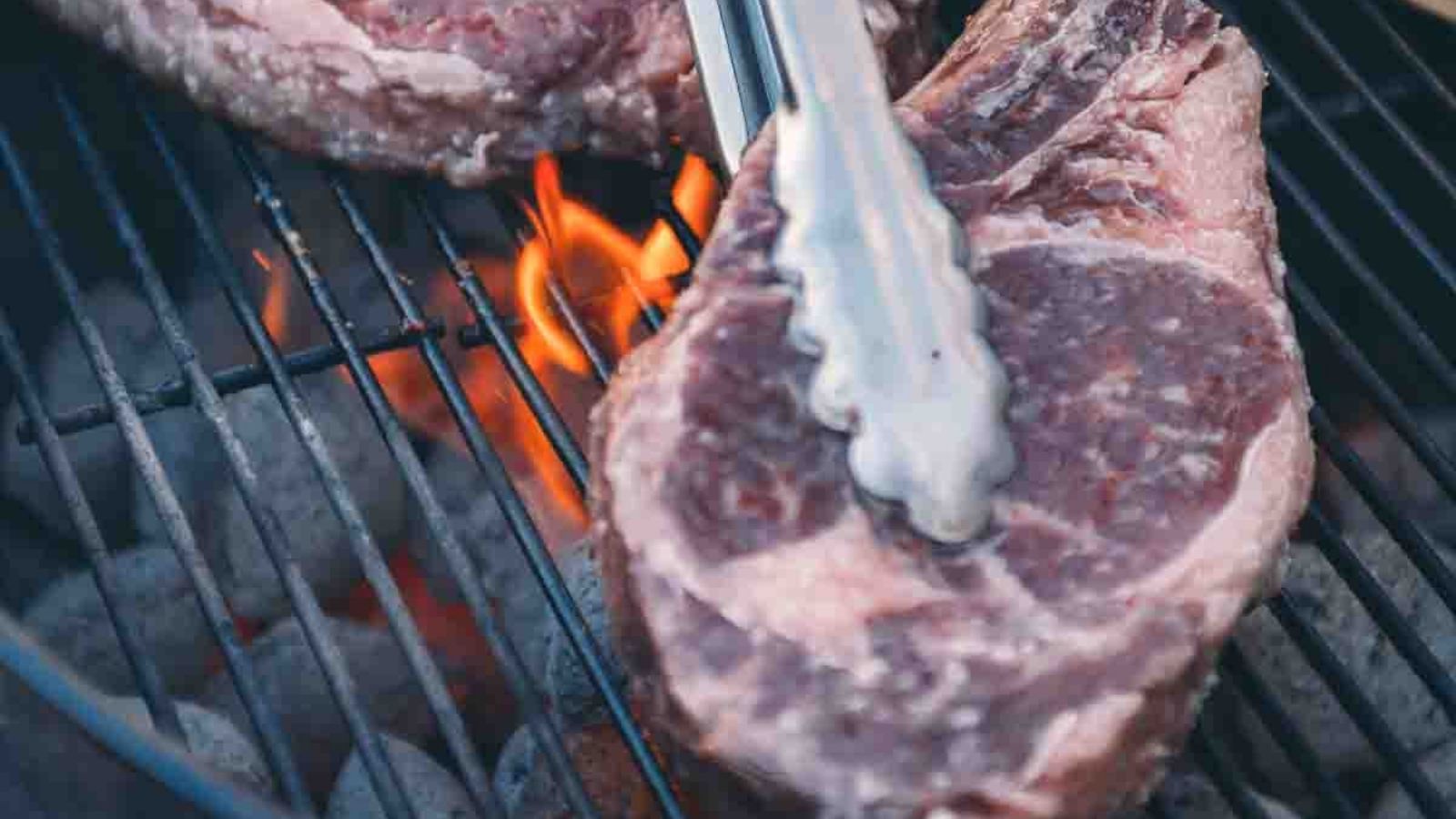 A raw steak on a grill with flames visible, being held by tongs.