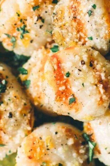 Close-up of cooked scallops garnished with herbs and black pepper, surrounded by lemon wedges and greens.