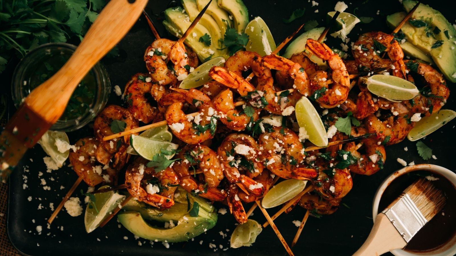 Grilled shrimp skewers garnished with lime wedges, sliced avocado, cilantro, and crumbled cheese on a black platter.