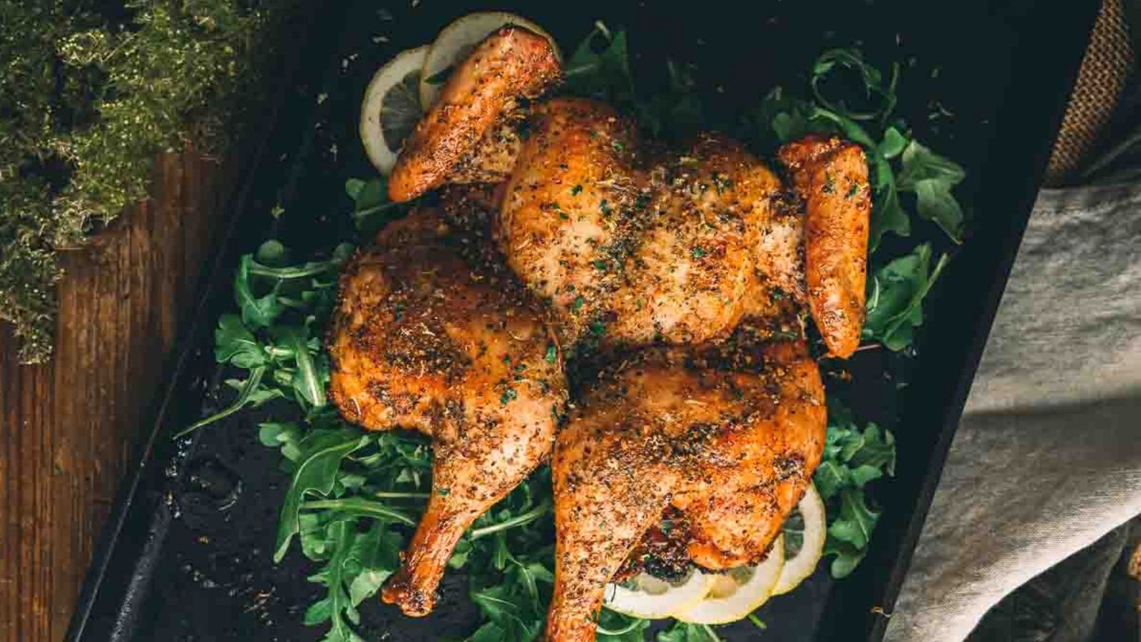 Spatchcock chicken with herbs on a bed of arugula and lemon slices, served on a dark tray.