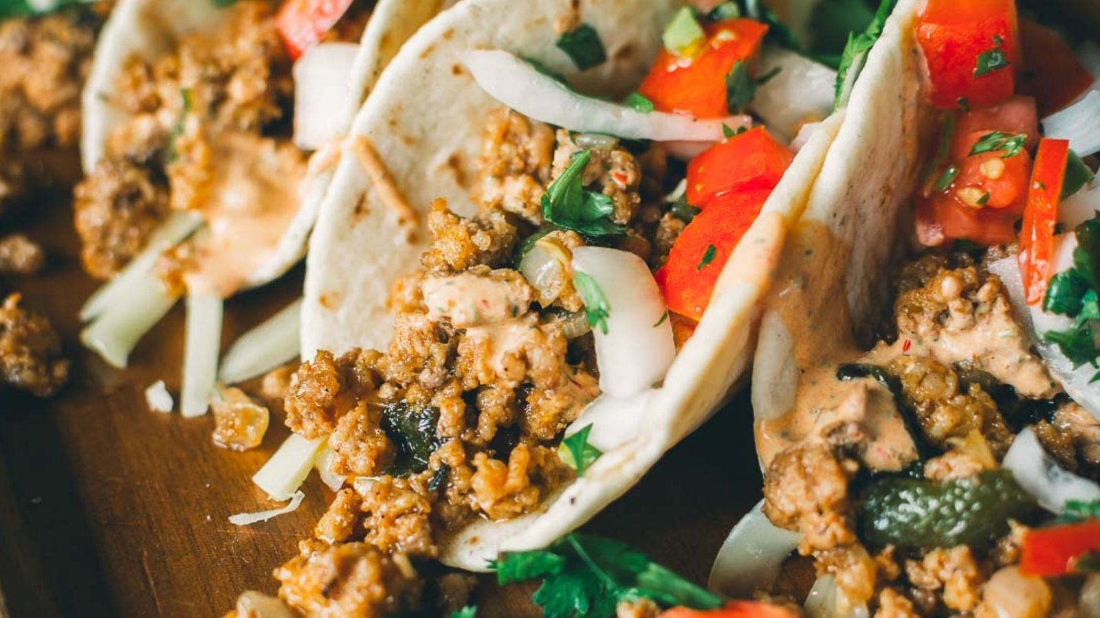 Three soft tacos filled with seasoned ground meat, chopped onions, tomatoes, and fresh cilantro on a wooden surface.