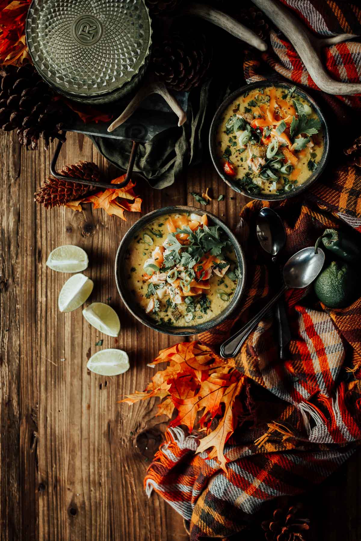 Two bowls of creamy soup on a wooden table, garnished with herbs and shredded vegetables. Surrounded by lime wedges, autumn leaves, spoons, and a plaid cloth.