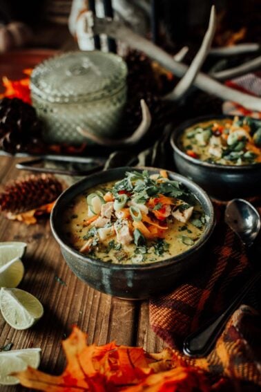 A bowl of creamy turkey soup garnished with green onions and herbs on a wooden table, surrounded by lime wedges, pinecones, and autumn leaves.