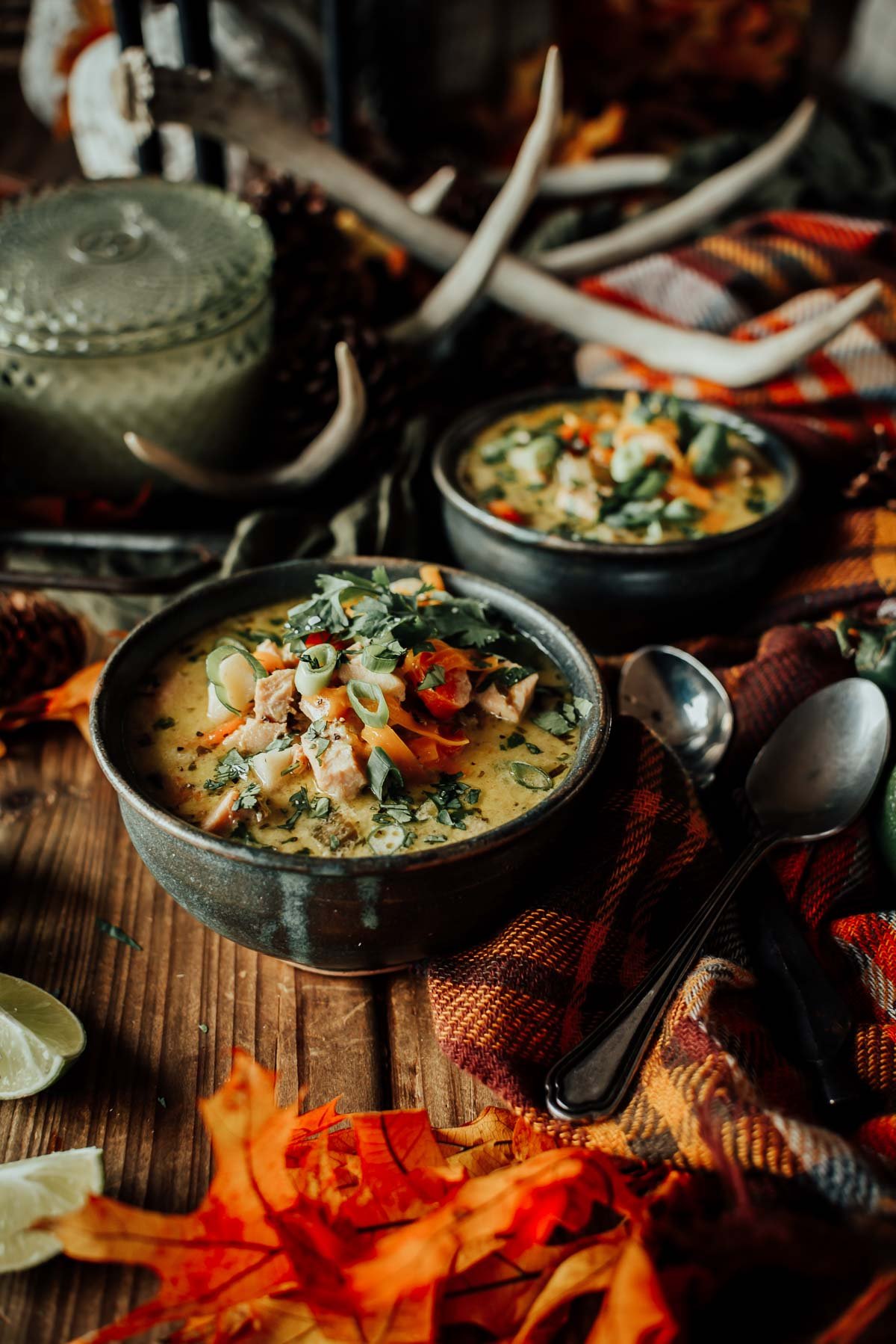 Two bowls of creamy turkey soup garnished with herbs and vegetables on a wooden table with autumn leaves, a plaid cloth, and cutlery.