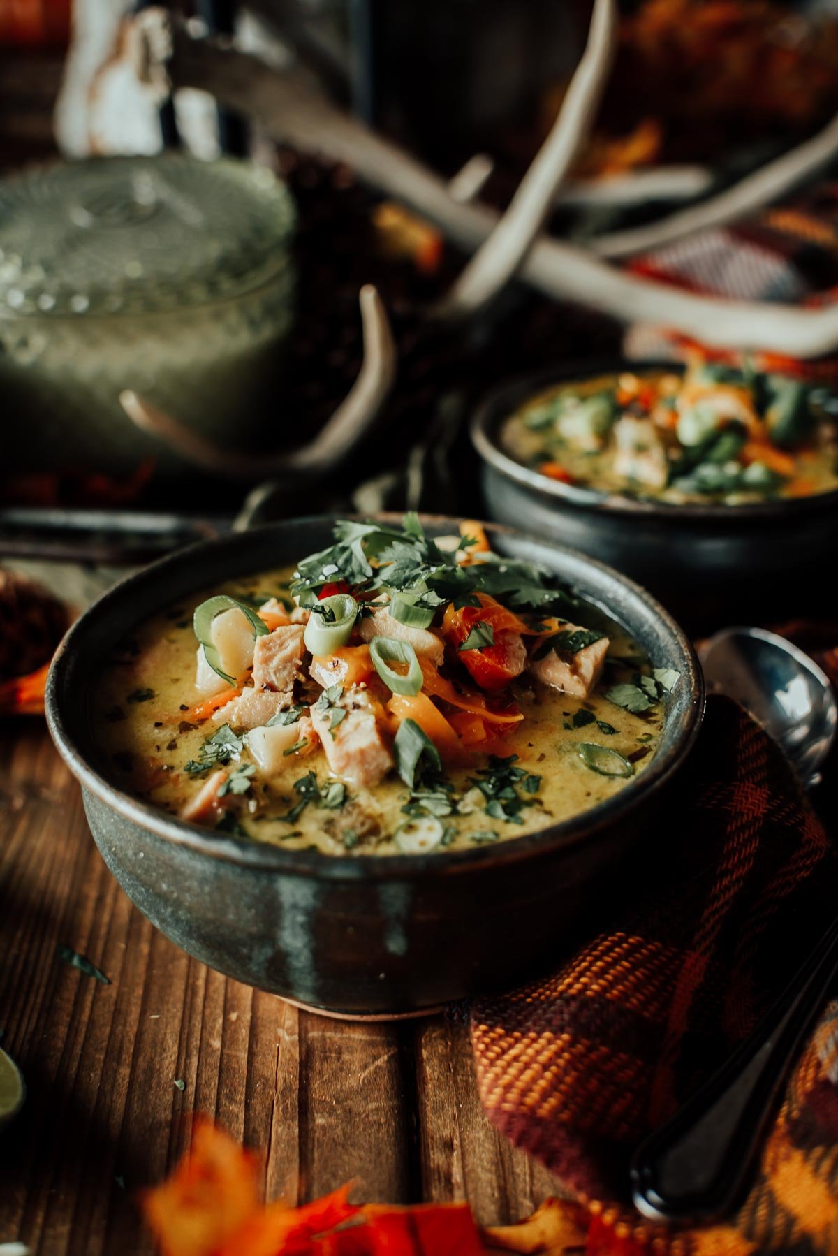 Bowl of creamy turkey soup garnished with sliced green onions and cilantro, set on a wooden table next to a plaid cloth and spoon.