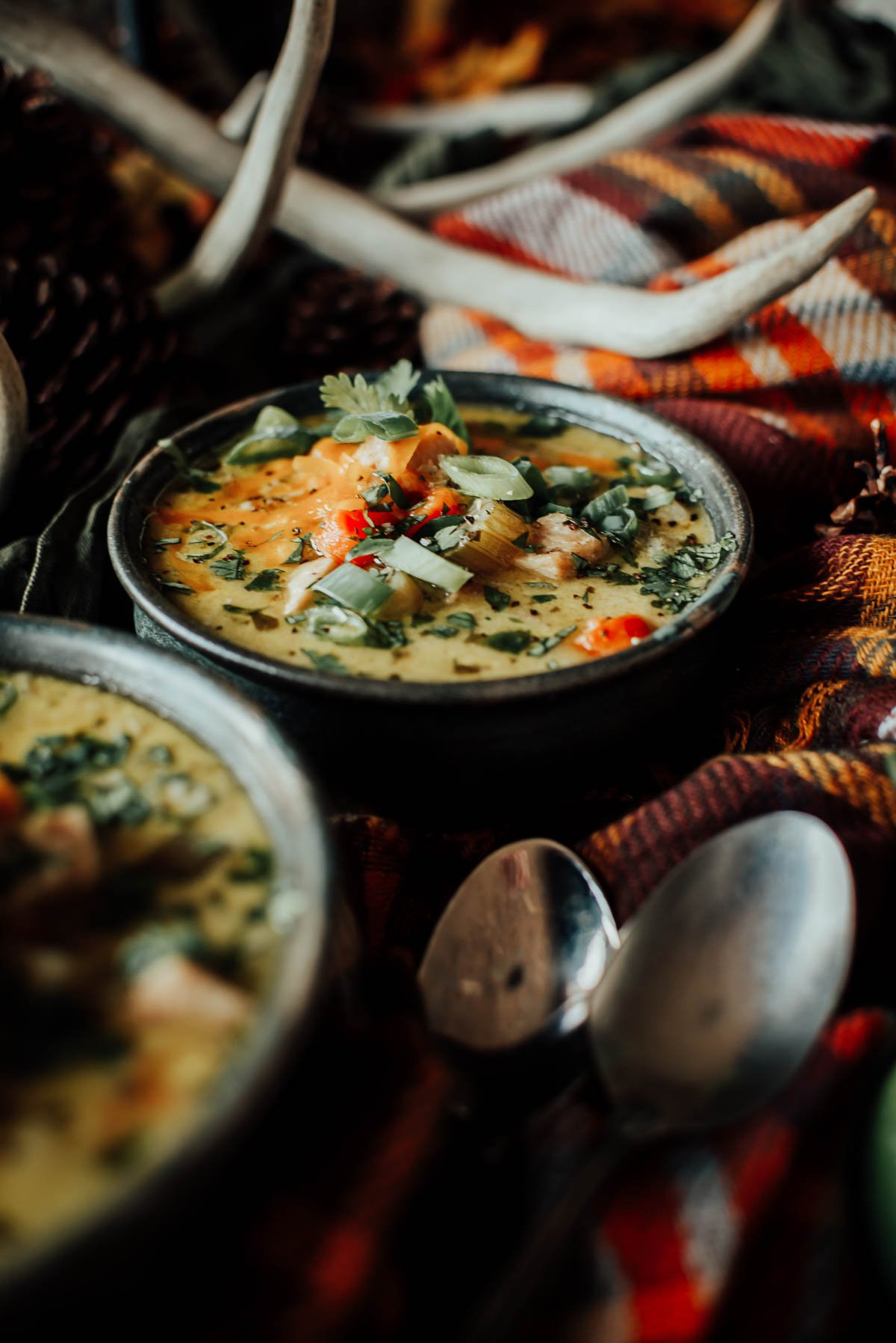 A bowl of creamy turkey soup topped with herbs and diced vegetables sits on a patterned fabric, with a spoon and decorative antlers nearby.