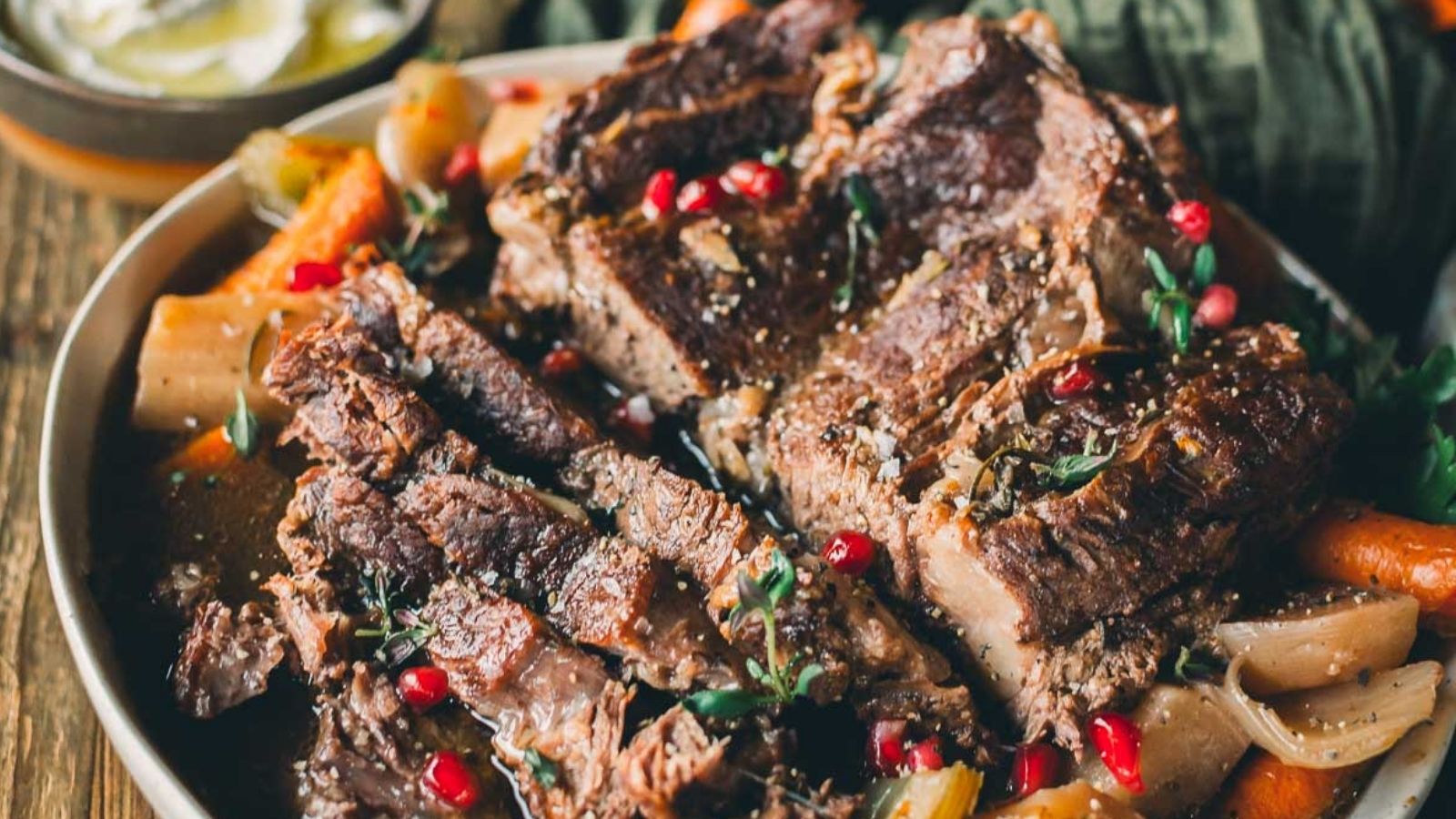 Close-up of sliced beef pot roast garnished with herbs and pomegranate seeds, served with carrots and onions.