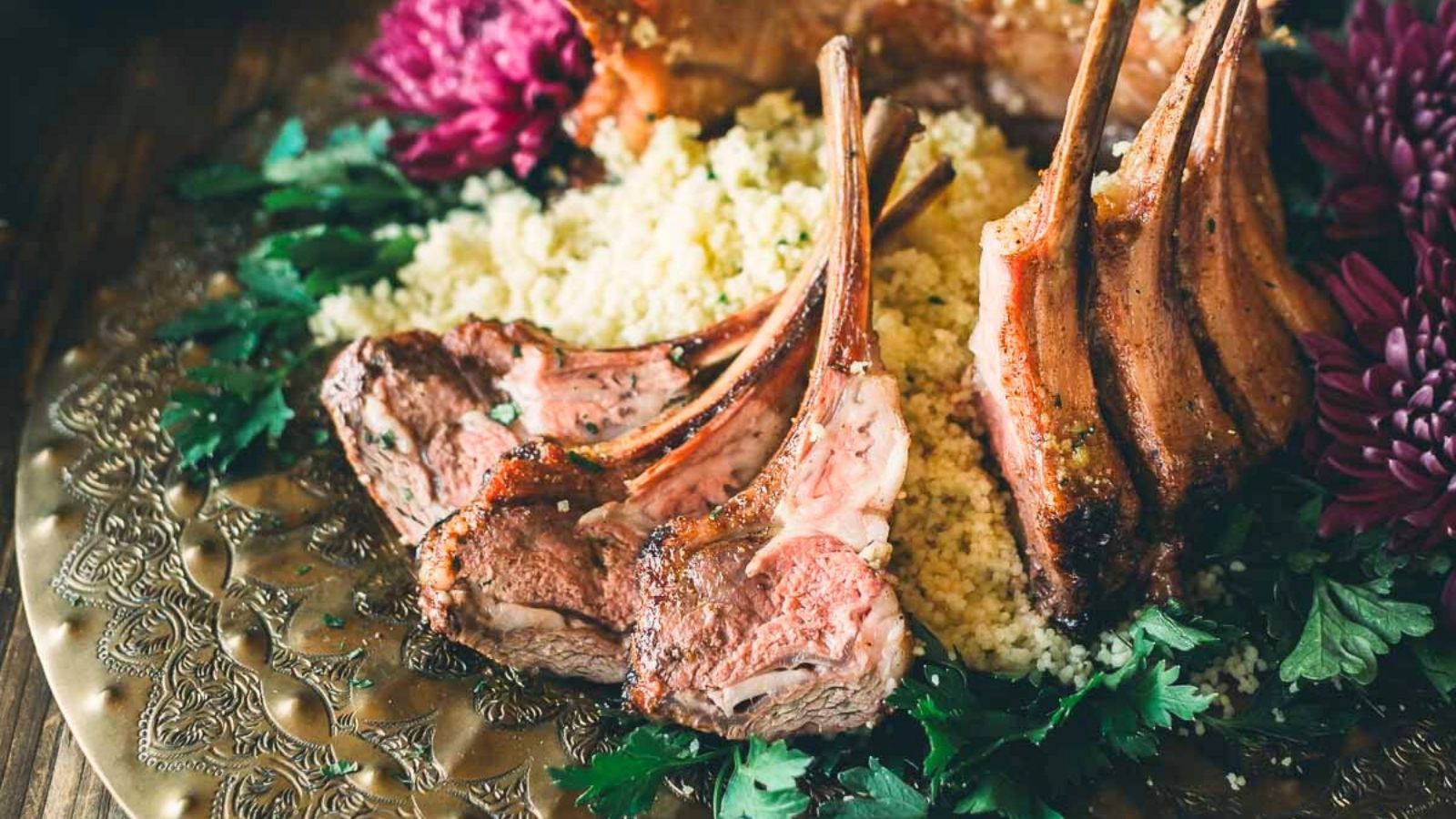 Grilled lamb chops with couscous and garnish on an ornate plate, surrounded by purple flowers and fresh herbs.