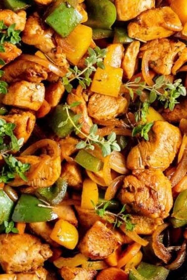 A skillet containing cooked chicken pieces mixed with colorful bell peppers, onions, and garnished with fresh herbs.