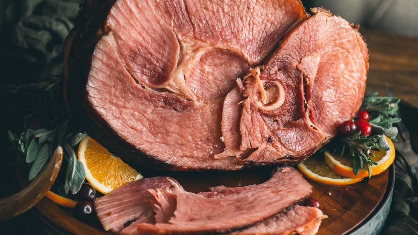 Close-up of a sliced glazed ham garnished with orange slices, fresh herbs, and berries on a wooden platter.