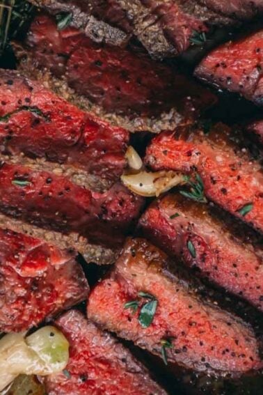 Sliced medium-rare steak on a board with herbs and garlic cloves, next to a large knife.