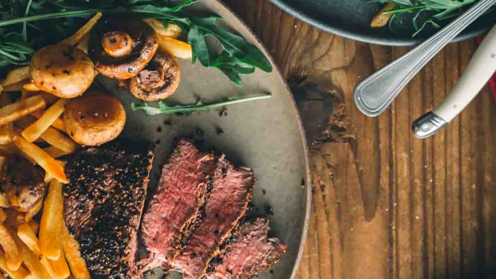 A plate with slices of beef, mushrooms, fries, and greens on a wooden table with cutlery nearby.