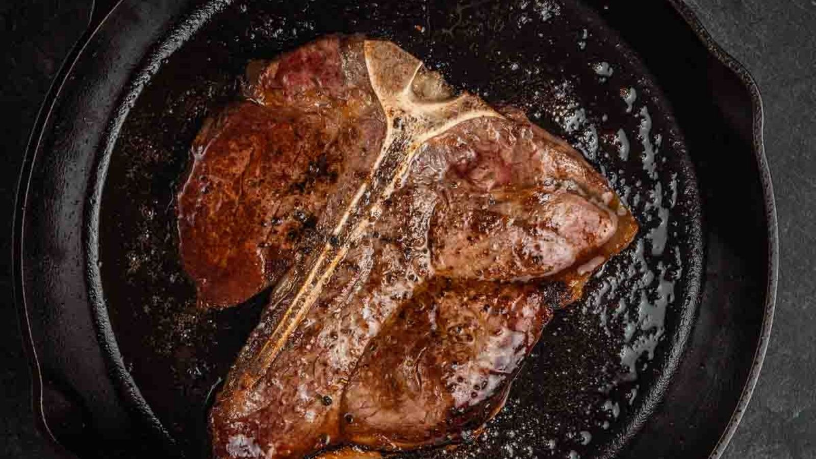 A cooked T-bone steak in a black skillet, seasoned and slightly charred.