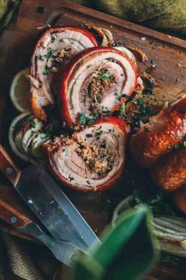 Sliced porchetta on a wooden board with herbs and spices, surrounded by knives and garnished with greens.