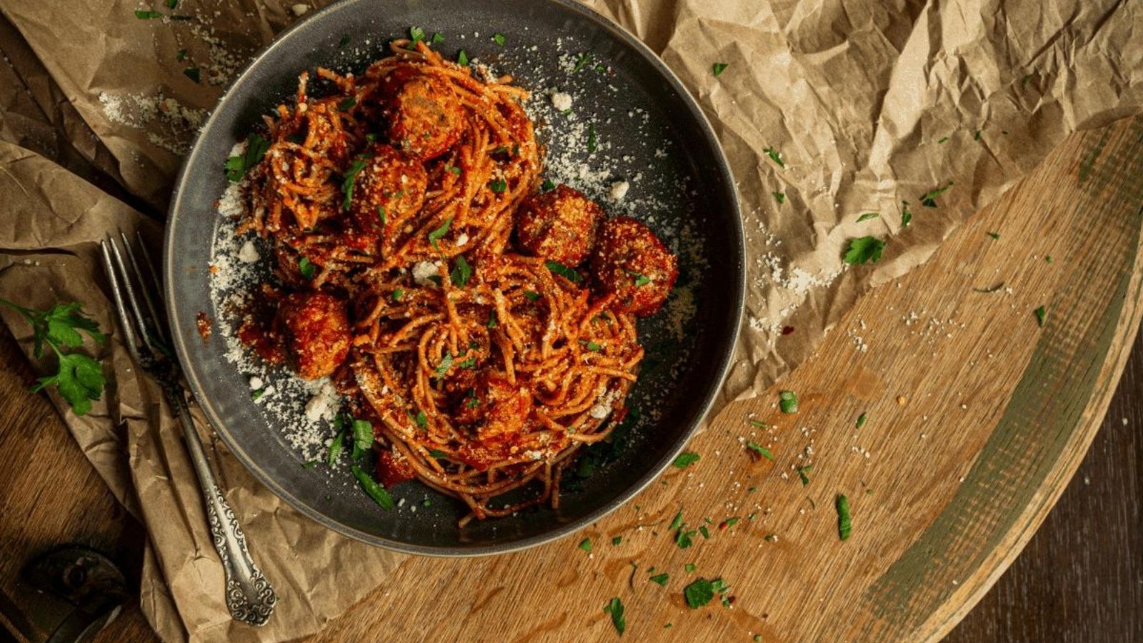 A plate of spaghetti with meatballs, garnished with grated cheese and parsley, on a wooden table covered with crumpled paper. A fork and spoon are placed beside the plate.