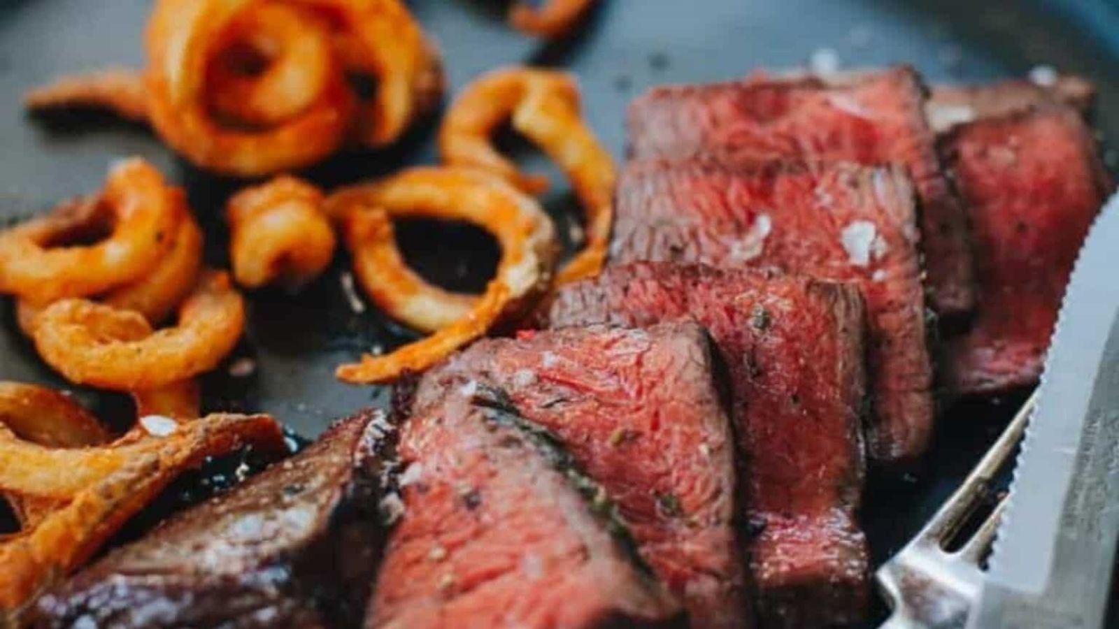 Slices of medium-rare steak on a plate with crispy curly fries and a knife.