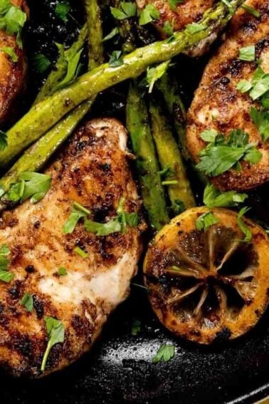 Grilled chicken breasts with asparagus, lemon slices, and herbs in a black skillet.