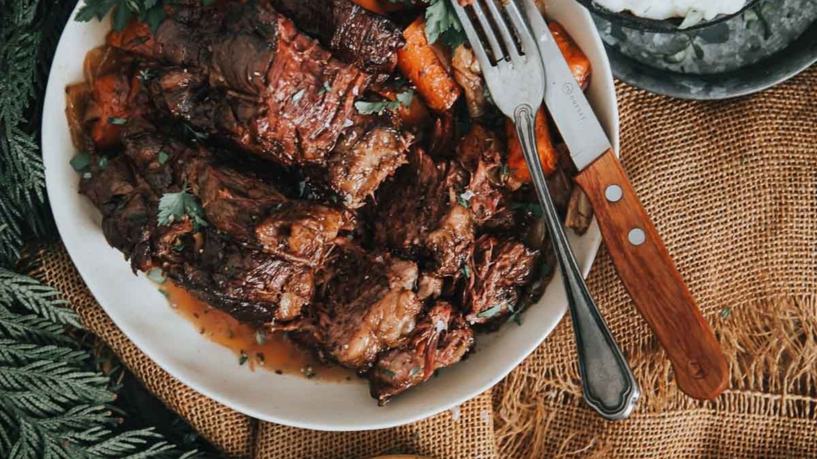 A plate of braised beef with carrots, garnished with herbs. Knife and fork rest on the plate, placed on a textured burlap tablecloth with greenery alongside.