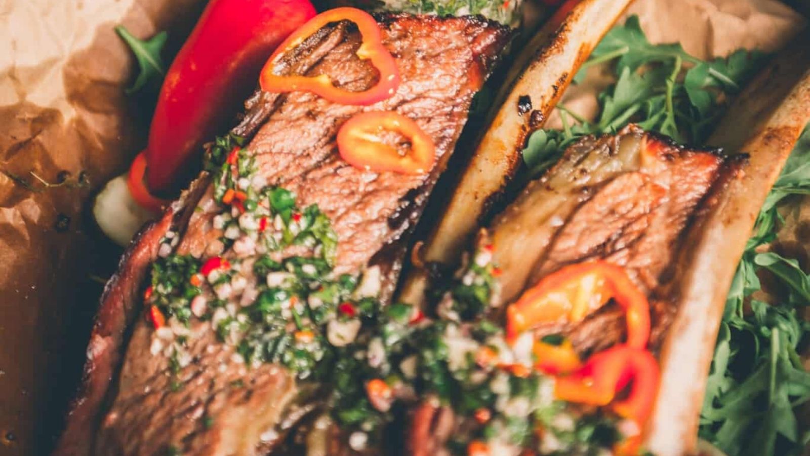 Grilled beef ribs garnished with green herbs and sliced red peppers on a bed of arugula.