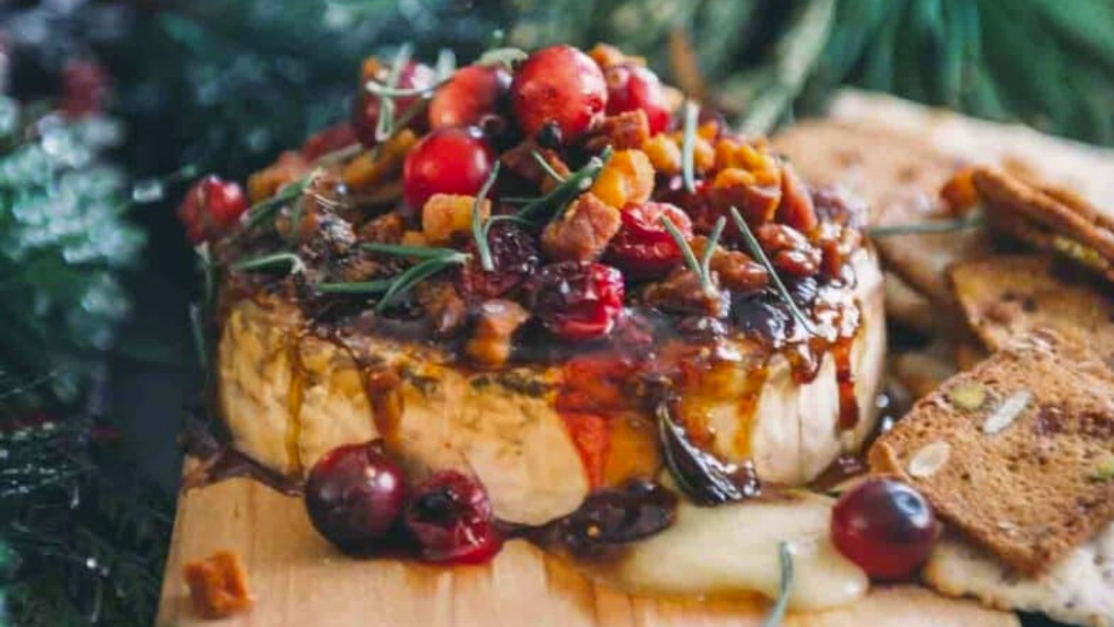 Brie cheese topped with cranberries, nuts, and herbs, served on a wooden board with crackers.