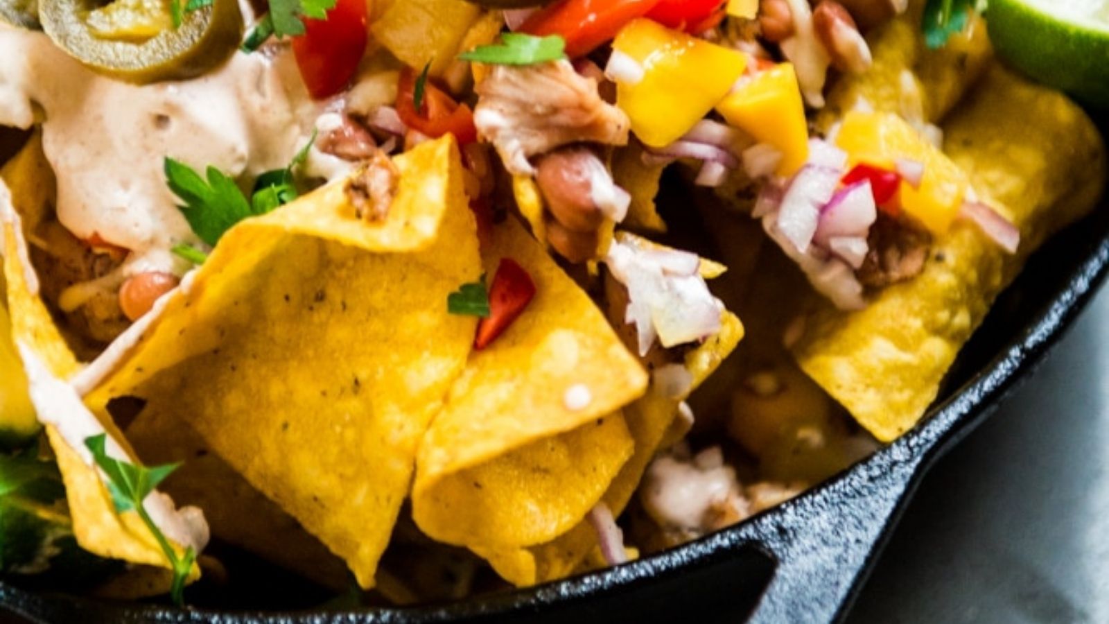 Close-up of loaded nachos topped with chopped onions, bell peppers, jalapeños, beans, and a creamy sauce in a black bowl.