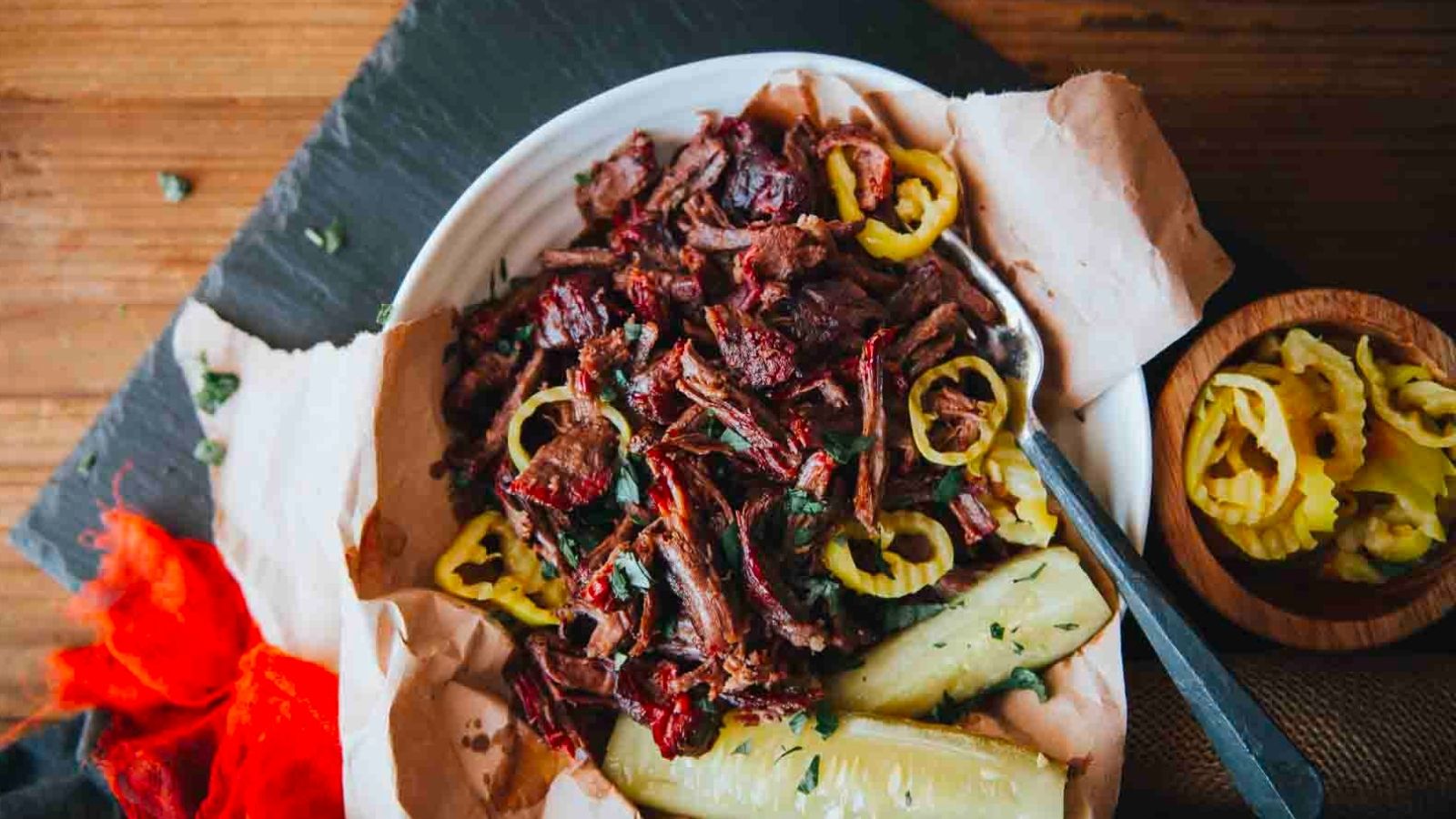 A plate of shredded beef with sliced yellow peppers and two dill pickles on the side, served on parchment paper.