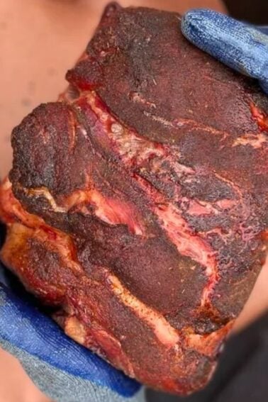 Gloved hands holding a large piece of seasoned, cooked meat on a brown surface.