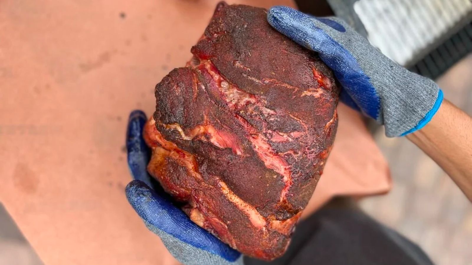 Gloved hands holding a large piece of seasoned, cooked meat on a brown surface.