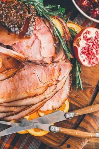Sliced roasted ham with garnish, served on a wooden board with knives and pomegranate halves. Nearby is a bowl of cranberry sauce on a checkered cloth.