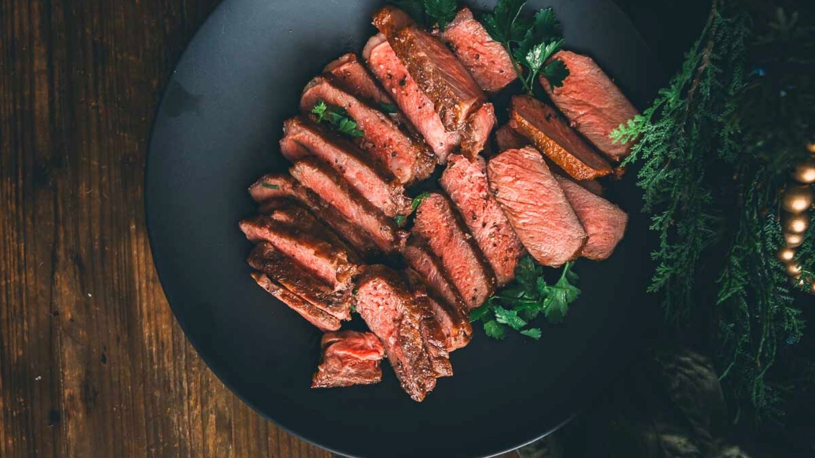 Slices of medium-rare beef steak arranged on a dark plate, garnished with fresh herbs, placed on a wooden table.