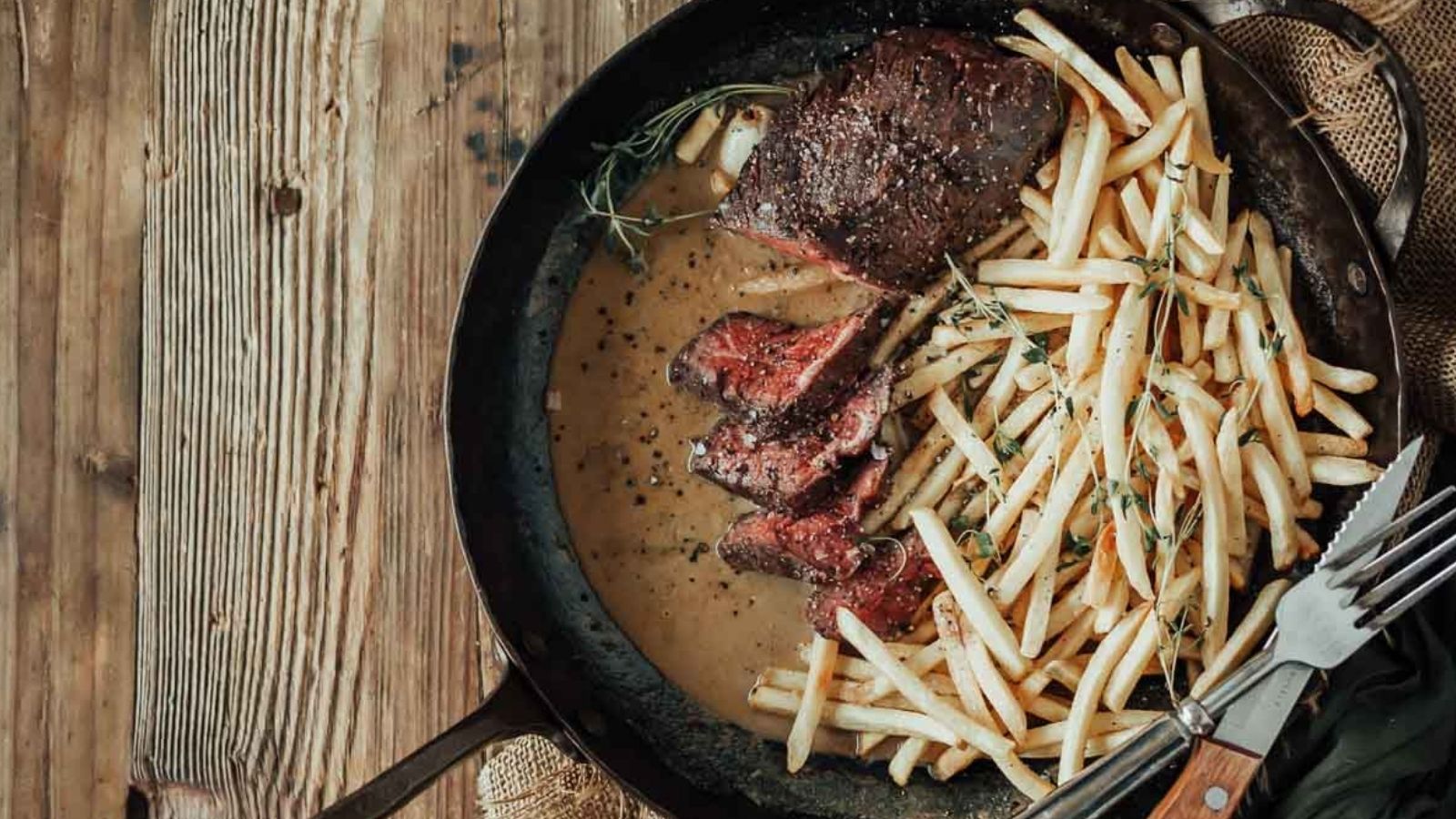 Steak slices and fries in a pan with sauce on a wooden table, accompanied by a fork and knife.